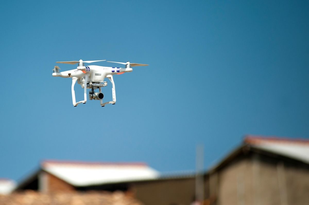 A white drone equipped with a camera flying in clear blue skies, with blurred buildings in the background.